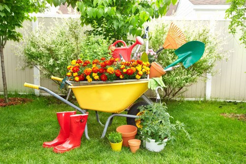 A garden scene featuring a yellow wheelbarrow filled with vibrant red and yellow flowers positioned on a well-maintained lawn in Plumstead. Surrounding the wheelbarrow are gardening tools including a red watering can, a green garden fork, and a blue plastic spade, indicating recent gardening activity. To the left, a pair of bright red rain boots rest on the grass, while nearby, several empty terracotta and plastic plant pots are arranged alongside a lush white flowering shrub in a pot. In the background, a white picket fence borders the garden, with mature trees and bushes providing privacy and shade. The outdoor space appears to be in good condition, with the grass evenly cut and the planting areas neatly maintained. Bright sunlight and clear weather create a cheerful atmosphere, making it an ideal environment for gardening services such as lawn care, planting, and outdoor maintenance, as offered by Lawn Mowing Plumstead. The scene emphasizes a tidy, inviting backyard suitable for garden enhancement and outdoor leisure in the Plumstead area, postcode SE18.