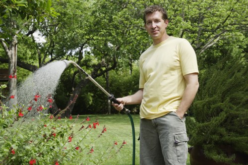 A man in a light yellow T-shirt and grey cargo shorts stands in a well-maintained garden in Plumstead, London, holding a garden hose with water spraying onto flowering plants. The garden features a lush, green lawn bordered by vibrant red flowers and various shrubs, with a backdrop of dense trees and bushes providing shade and privacy. The ground surface includes neatly trimmed grass, and the scene appears to be set during a bright, partly cloudy day with natural sunlight illuminating the outdoor space. The overall environment suggests a cared-for landscaped garden, suitable for outdoor leisure and gardening services offered by Lawn Mowing Plumstead, with the scene emphasizing lawn and garden maintenance activities typical of local gardening and landscaping expertise.